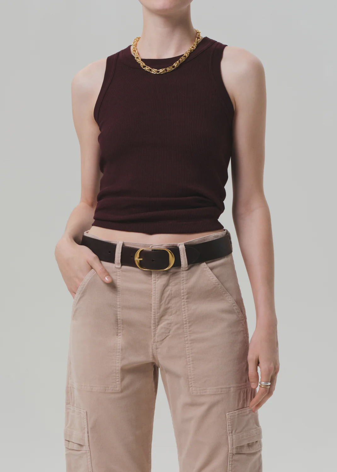 A person wears the Isabel Rib Tank F25 by Citizens Of Humanity/AGOLDE—a sleeveless, high-neck dark burgundy ribbed top—paired with beige cargo pants, a black belt with gold buckle, gold chain necklace, and rings. Their face is not visible.