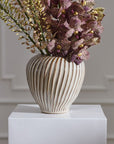 Tomar Vase from Accent Decor with purple orchids and greenery on a white pedestal against a pale wall.