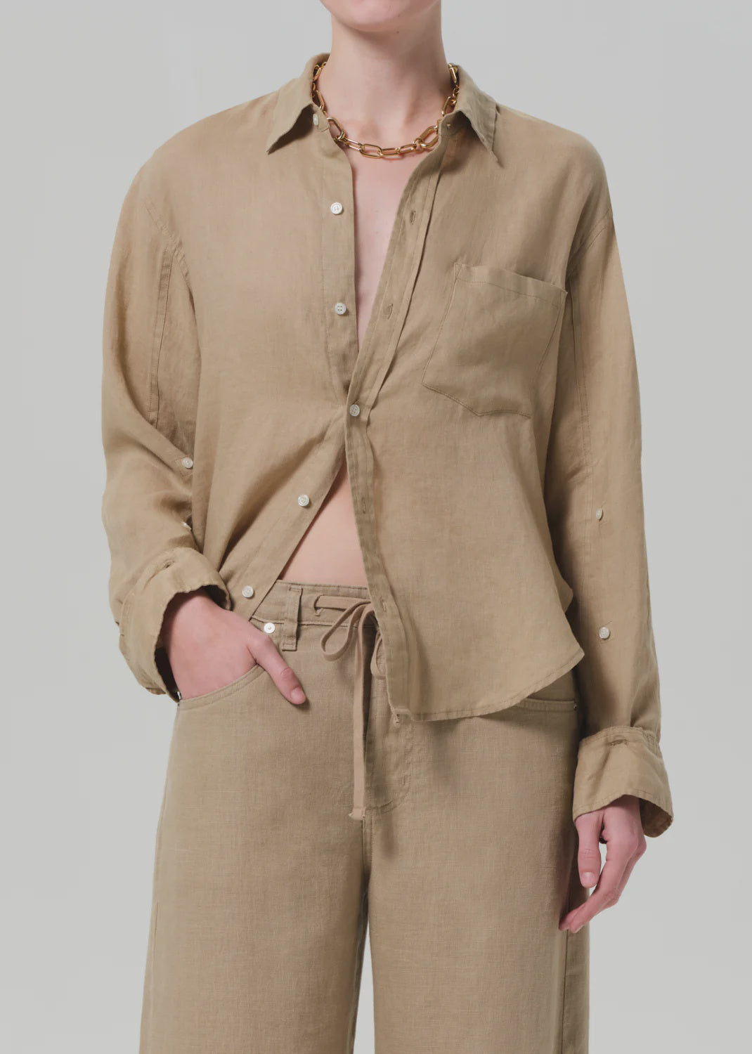 Wearing the Kayla Shirt F25 by Citizens Of Humanity/AGOLDE, a breathable, lightweight beige linen collared shirt with rolled sleeves and a partially unbuttoned, tucked-in look, paired with matching drawstring pants and a chunky gold chain necklace.
