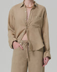 Wearing the Kayla Shirt F25 by Citizens Of Humanity/AGOLDE, a breathable, lightweight beige linen collared shirt with rolled sleeves and a partially unbuttoned, tucked-in look, paired with matching drawstring pants and a chunky gold chain necklace.