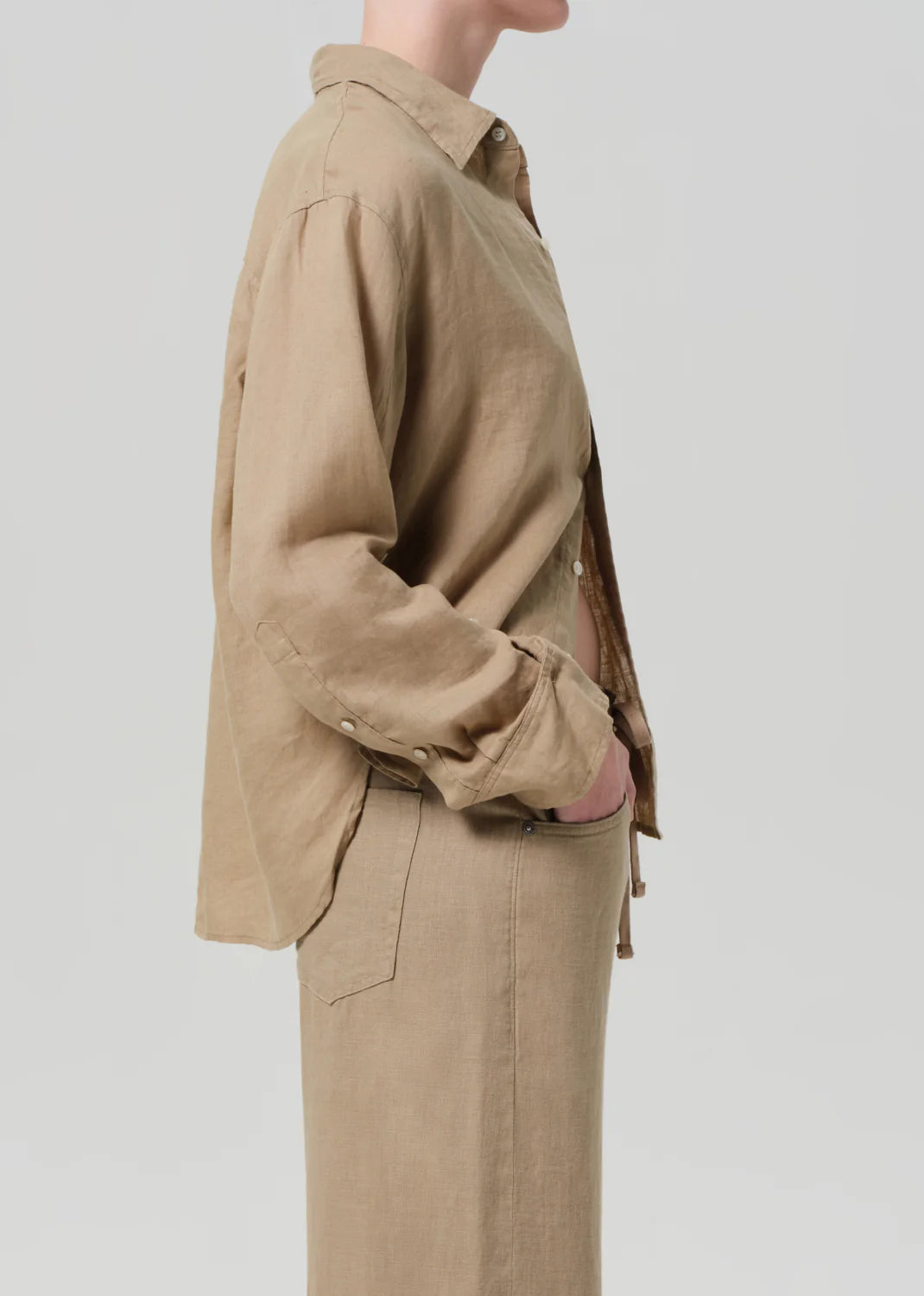 A person stands sideways with one hand in their pocket, wearing the Kayla Shirt F25 by Citizens Of Humanity/AGOLDE—a beige linen shirt with rolled-up sleeves and matching pants—exuding relaxed style against a light gray background.