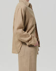 A person stands sideways with one hand in their pocket, wearing the Kayla Shirt F25 by Citizens Of Humanity/AGOLDE—a beige linen shirt with rolled-up sleeves and matching pants—exuding relaxed style against a light gray background.