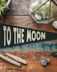 The Faire To The Moon Felt Pennant, featuring large cream letters, sits on a rustic table amid plants and crystals—ideal as vintage nursery decor or a charming family love banner.