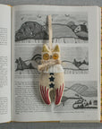 The Rocket Cat Elton John, a handmade ornament filled with cotton and adorned with star patterns and a ribbon, rests on an open book featuring hand-illustrated black and white nature illustrations. The book lies on a textured gray surface, showcased by the brand Faire.