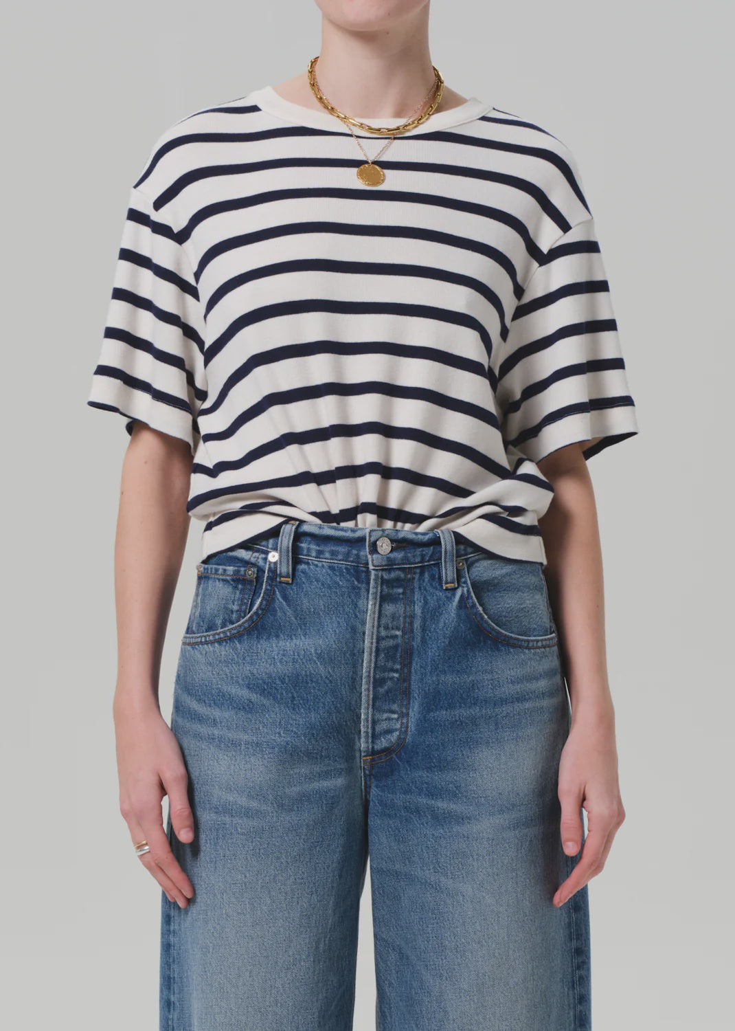 A person is wearing the Goldie Tee, a lightweight rib cotton blend from Citizens Of Humanity/AGOLDE, knotted at the waist with high-waisted blue jeans. They accessorize with a gold necklace and ring against a plain light-colored background.