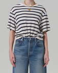 A person is wearing the Goldie Tee, a lightweight rib cotton blend from Citizens Of Humanity/AGOLDE, knotted at the waist with high-waisted blue jeans. They accessorize with a gold necklace and ring against a plain light-colored background.