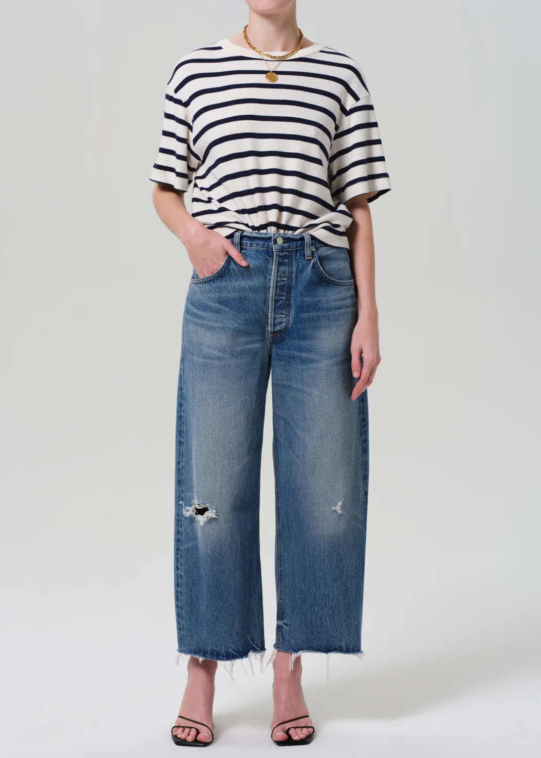 Dressed in the Citizens Of Humanity/AGOLDE Goldie Tee, an oversized black and white striped rib cotton top tucked into high-waisted wide-leg distressed jeans, the person completes their look with black strappy heels and a gold necklace against a plain gray background.