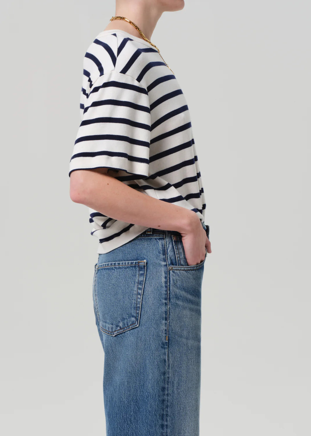 A model in profile wears the Goldie Tee, an oversized black and white striped shirt by Citizens Of Humanity/AGOLDE, tucked into blue jeans with their right hand in their pocket against a plain background.