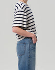 A model in profile wears the Goldie Tee, an oversized black and white striped shirt by Citizens Of Humanity/AGOLDE, tucked into blue jeans with their right hand in their pocket against a plain background.