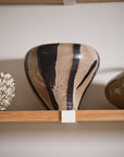 The Atwood Vase by Accent Decor—a hand painted ceramic piece with a beige base and bold vertical black brushstrokes—rests on a wooden shelf, surrounded by sculptural vessels and decorative objects.