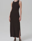 A person wears the Isabel Tank Dress by Citizens Of Humanity/AGOLDE—a sleeveless black midi with side slit—styled with a chunky chain necklace and black sandals, standing against a plain, light-colored background.