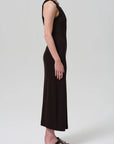 A person stands in profile against a plain background, wearing the Citizens Of Humanity Isabel Tank Dress—an ankle-length, sleeveless black ribbed dress—paired with black sandals, a chunky necklace, and rings on one hand.