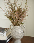 The Lines Vase by Accent Decor, a striped ceramic piece with two handles, displays long-stemmed greenery and white flowers. Handcrafted, it rests on a wooden surface beside stacked books.
