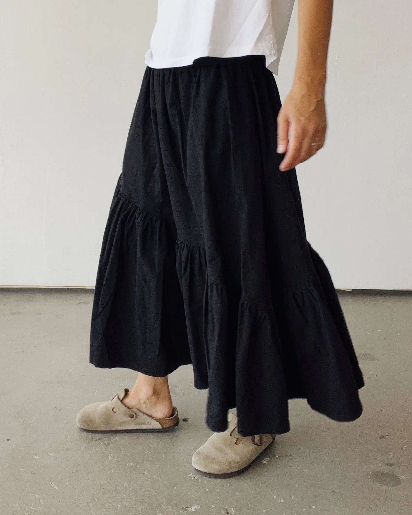A person wearing a white t-shirt, Hey Gang’s The Ranch Skirt in black with ruffles, and beige slip-on clogs stands on a gray floor against a plain light-colored wall.