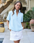 A woman stands in a courtyard with plants and a tree, wearing the Patty Popover SS Henley by Frank & Eileen and a white skirt. Her hands are in her pockets, with a basket visible in the background.