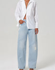 A person in a Polina Shirt from Citizens Of Humanity/AGOLDE, paired with light blue high-waisted wide-leg jeans featuring a diagonal pattern and heeled sandals. One hand is in their pocket as they stand against a plain background.