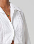 A person wearing the Polina Shirt, a sleek white collared piece with pleated tailoring details and an open collar by Citizens Of Humanity/AGOLDE. They complement it with a gold necklace featuring a green gemstone pendant against a plain, light gray backdrop.