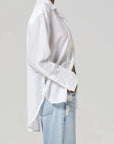 A model in a Polina Shirt by Citizens Of Humanity/AGOLDE, featuring fine tailoring, pairs it with light blue ripped jeans. The plain background highlights only their torso and legs as they stand sideways with hands in pockets.