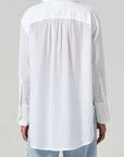 Back view of a person in the Polina Shirt by Citizens Of Humanity/AGOLDE, featuring a white loose-fitting long-sleeve collared design in a cotton silk blend, paired with light blue jeans. A pleated detail near the shoulders is highlighted against a neutral gray background. Made in India.
