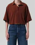 A person wears the Lowen Polo Shirt by Citizens Of Humanity/AGOLDE in brown supima cotton, styled oversized with high-waisted dark blue jeans. Gold necklace and rings complete the look against a plain light background.
