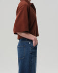 A person stands in profile with one hand in their pocket, wearing the oversized brown Lowen Polo Shirt by Citizens Of Humanity/AGOLDE and blue high-waisted jeans against a plain light background.
