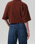 A person faces away, wearing a loose-fitting brown Lowen Polo Shirt by Citizens Of Humanity/AGOLDE with elbow-length sleeves and high-waisted blue jeans with visible back pockets, set against a neutral light gray background.