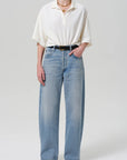 A person wears the Citizens Of Humanity/AGOLDE Lowen oversized white short-sleeve polo in breathable pique, paired with light blue high-waisted wide-leg jeans, a black belt, black heeled sandals, and layered gold necklaces against a plain light gray background.