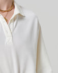 A person wears the cream Lowen Polo Shirt by Citizens Of Humanity/AGOLDE, featuring breathable pique fabric and buttons, styled with layered gold chain necklaces against a neutral backdrop; only their upper torso, neck, and shoulder are visible.