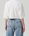 Someone is seen from the back against a plain light background, wearing Citizens Of Humanity/AGOLDE's Lowen Polo Shirt—a loose-fitting, cropped white pique top with elbow-length sleeves—paired with high-waisted, light blue jeans.