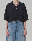 A person wears the Citizens Of Humanity/AGOLDE Lowen Polo Shirt, an oversized black pique top, tucked into high-waisted wide-leg blue jeans with front patch pockets, styled with a chunky silver chain necklace and ring against a plain light background.