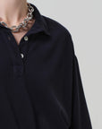 A person wears the Citizens Of Humanity/AGOLDE Lowen Polo Shirt in dark pique fabric, paired with a chunky silver chain necklace, focusing on the upper chest and neck against a plain, light background.