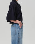 Against a plain light background, a person stands in profile with hands in pockets, wearing the Lowen Polo Shirt by Citizens Of Humanity/AGOLDE and high-waisted light blue jeans.