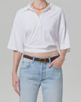 Wearing a white lowen polo shirt by Citizens Of Humanity/AGOLDE, light blue high-waisted jeans with a brown belt, and gold jewelry, a person stands with one hand in their pocket against a plain background.