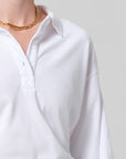 A person wears the Citizens Of Humanity/AGOLDE lowen polo shirt, a white short sleeve polo with buttons, paired with a gold chain necklace, shown from shoulders to mid-chest against a plain gray background.