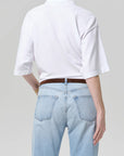 A person is seen from the back wearing a plain white Citizens Of Humanity/AGOLDE lowen polo shirt tucked into light blue high-waisted jeans with a brown belt, against a plain light gray background.