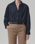A person wears the Ana Cropped Shirt in Piave by Citizens Of Humanity/AGOLDE, a dark indigo Japanese denim button-up, tucked into high-waisted tan trousers with a black belt, one hand in their pocket and face not visible.