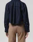 A person wears the Ana Cropped Shirt in Piave by Citizens Of Humanity/AGOLDE, a dark blue Japanese denim jacket with long sleeves, paired with high-waisted tan pants, seen from the back against a plain light background.