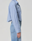 A person stands in profile wearing the ana cropped shirt in banker blue plaid by Citizens Of Humanity/AGOLDE and high-waisted light blue jeans against a plain light background.