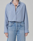 A person wears the ana cropped shirt in banker blue plaid by Citizens Of Humanity/AGOLDE, tucked into high-waisted light blue jeans, with rolled sleeves, a black pendant necklace, and rings, standing against a plain background.
