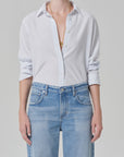 A person wears the Citizens Of Humanity/AGOLDE avni slim shirt in blue micro stripe with rolled sleeves and layered gold necklaces, paired with high-waisted light blue jeans. The background is plain light gray.