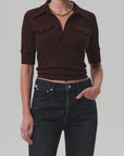 Wearing the Amina Polo Shirt by Citizens Of Humanity/AGOLDE—a dark burgundy, ribbed cotton short-sleeve collared top with buttons—paired with high-waisted black jeans and a gold chain necklace against a plain background.