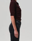A person stands in profile against a plain background, wearing the Amina Polo Shirt by Citizens Of Humanity/AGOLDE— a ribbed cotton, short-sleeve burgundy top with collar and pocket—paired with high-waisted black jeans. Their left arm is relaxed by their side.