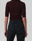 A person stands with their back to the camera, wearing the Amina Polo Shirt by Citizens Of Humanity/AGOLDE in dark burgundy ribbed cotton and high-waisted black denim jeans against a plain, light background.