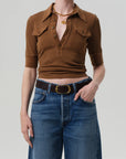 A person wears the Citizens Of Humanity/AGOLDE Amina Polo Shirt—ribbed cotton, brown with front patch pockets—tucked into high-waisted blue jeans with a black belt and gold buckle, finished with layered gold necklaces and a ring.