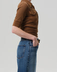 A person in the Citizens Of Humanity/AGOLDE Amina Polo Shirt and blue jeans with front patch pockets stands in profile with one hand in their pocket against a plain light gray background.