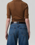 A person is seen from the back wearing the ribbed cotton Amina Polo Shirt by Citizens Of Humanity/AGOLDE in brown, paired with high-waisted blue jeans with large back pockets, against a plain, light background.