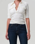 A person wears the Citizens Of Humanity/AGOLDE Amina Polo Shirt—a fitted, white ribbed cotton knit with a deep V-neck, button details, and chest pockets—paired with dark blue jeans and a necklace against a plain light background.