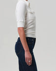 A person in profile wears the Amina Polo Shirt by Citizens Of Humanity/AGOLDE, a white ribbed cotton, short-sleeve top with a collar and front pocket detail, paired with dark blue jeans against a plain light background.