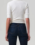 A person is shown from the back, wearing the Amina Polo Shirt by Citizens Of Humanity/AGOLDE in white, styled with dark blue jeans with front pockets. The background is plain and light gray.