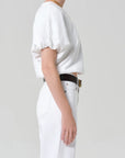A person in profile wears the Citizens Of Humanity/AGOLDE gia bubble tee with puff sleeves, white high-waisted pants, a black belt with gold buckle, a gold chain necklace, and a ring against a plain light grey background.
